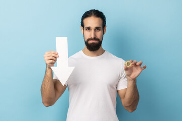 Portrait of sad unhappy man with beard wearing white T-shirt showing gold bitcoin and arrow pointing down, downgrade of electronic currency. Indoor studio shot isolated on blue background.