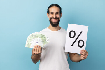 Portrait of positive satisfied man with beard wearing white T-shirt holding paper with percent sign...