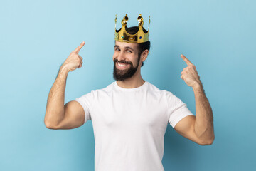 Portrait of satisfied happy man with beard wearing white T-shirt pointing fingers on golden crown on his head, showing his authority. Indoor studio shot isolated on blue background. - obrazy, fototapety, plakaty