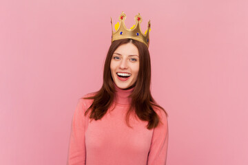 Happy confident smiling woman with brown hair standing in gold crown, looking at camera with positive expression, laughing, wearing rose turtleneck. Indoor studio shot isolated on pink background - obrazy, fototapety, plakaty