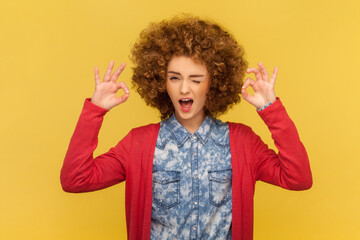 Portrait of playful cheerful woman with Afro hairstyle winking to camera, showing okay gesture with both hands, saying all is ok. Indoor studio shot isolated on yellow background. - obrazy, fototapety, plakaty