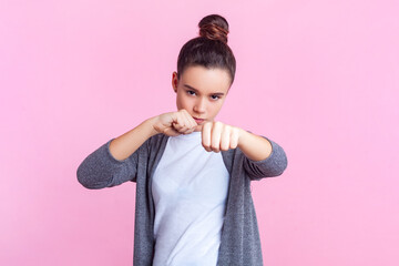 Portrait of strict angry teenage girl with bun hairstyle in casual clothes standing with clenched fists being ready to attack. Indoor studio shot isolated on pink background.