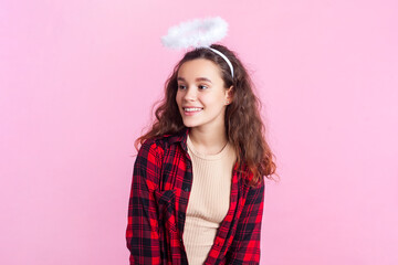 Portrait of charming teenage girl with wavy hair in red checkered shirt and nimb over head looking away with dreamy expression. Indoor studio shot isolated on pink background.