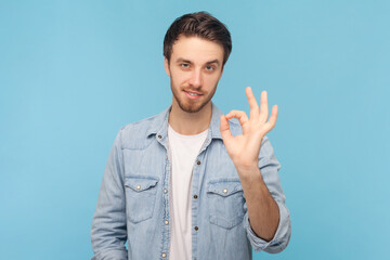 Okay, its fine. Portrait of unshaven man says no problem, shows excellent gesture, gives recommendation, demonstrates positive approval, wearing denim shirt. Indoor shot isolated on blue background.