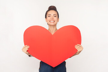 Portrait of romantic cheerful woman with hair bun holding big red heart, looking at camera with toothy smile, expressing love, wearing denim overalls. Indoor studio shot isolated on white background