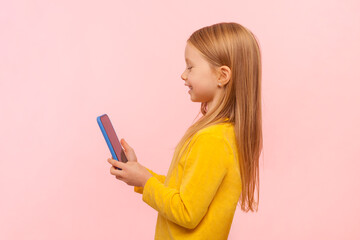 Naklejka premium Side view portrait of adorable cute blonde little girl using cell phone with happy expression, playing games, enjoying, wearing yellow jumper. Indoor studio shot isolated on pink background.