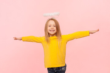 Portrait of smiling adorable blonde little girl with nimb over head standing with spread arms, being happy and in good mood, wearing yellow jumper. Indoor studio shot isolated on pink background.