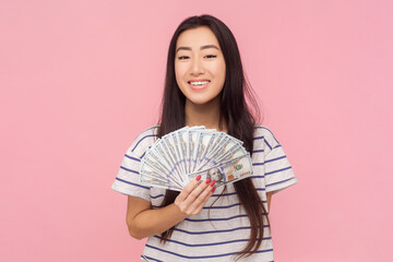 Portrait of satisfied pride rich woman with long brunette hair holding fan of dollar banknotes, wearing striped T-shirt. Indoor studio shot isolated on pink background.