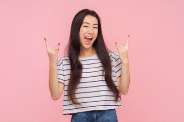 Portrait of crazy excited amazed woman with long brunette hair having fun on festival showing rock and roll gesture, wearing striped T-shirt. Indoor studio shot isolated on pink background.