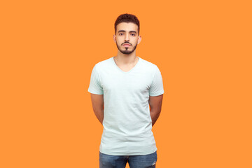 Fototapeta na wymiar Portrait of serious handsome young bearded man wearing T-shirt standing looking at camera with bossy facial expression. Indoor studio shot isolated on orange background.