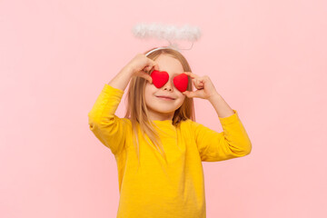 Portrait of angelic blonde little girl with nimb over head covering her eyes with small red hearts, smiling friendly, wearing yellow jumper. Indoor studio shot isolated on pink background.
