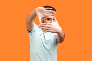 Portrait of concentrated focused young bearded man wearing T-shirt looking at camera through frame of fingers taking picture. Indoor studio shot isolated on orange background.