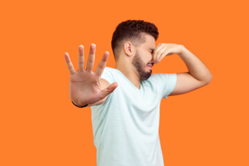 Portrait of displeased young bearded man wearing T-shirt pinching his nose turning away feeling disgust bad smell. Indoor studio shot isolated on orange background. - obrazy, fototapety, plakaty