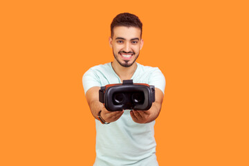 Portrait of handsome pleased young bearded man wearing T-shirt holding out virtual reality headset...