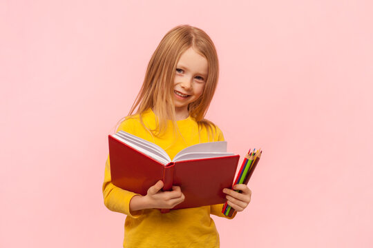 Portrait of charming blonde little girl reading book and holding colorful pencils, looking smiling at camera, wearing yellow jumper. Indoor studio shot isolated on pink background.