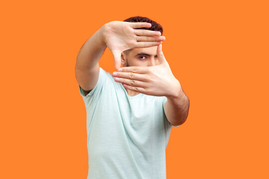 Portrait of concentrated focused young bearded man wearing T-shirt looking at camera through frame of fingers taking picture. Indoor studio shot isolated on orange background.