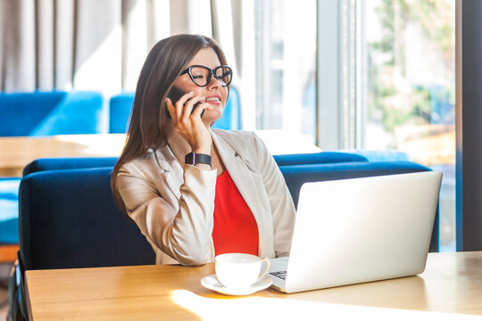 Portrait of confident woman working on laptop and talking on smartphone with business partners positive emotions, wearing jacket and re shirt. Indoor shot, cafe or office background.