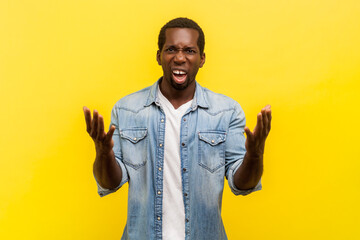 Portrait of angry annoyed man standing with raised arms and frowning face expressing anger and aggression, wearing denim casual shirt. Indoor studio shot isolated on yellow background. - obrazy, fototapety, plakaty