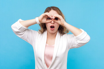 Portrait of shocked astonished blond woman with wavy hair standing raising arms showing binocular gesture saying wow, wearing white shirt. Indoor studio shot isolated on blue background.