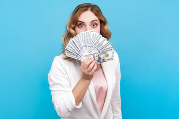 Portrait of shocked woman with wavy hair covering half of face with fan of money standing with her...