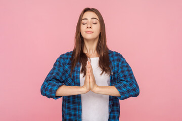 Portrait of calm concentrated brown haired woman standing with palms together keeps eyes closed practicing yoga, wearing checkered shirt. Indoor studio shot isolated on pink background.