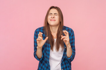 Portrait of hopeful brown haired woman standing with closed eyes and crossed fingers making wish dreaming, wearing checkered shirt. Indoor studio shot isolated on pink background.
