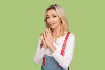 Portrait of envy cunning beautiful adult blond woman presses fingers together, planning sly prank, looking at camera, wearing denim overalls. Indoor studio shot isolated on light green background - obrazy, fototapety, plakaty