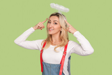 Portrait of delighted cheerful angelic adult blond woman pointing at her nimb over head, looking smiling at camera, wearing denim overalls. Indoor studio shot isolated on light green background