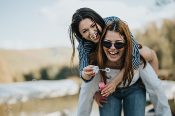 Two happy young women enjoying a playful moment with soap bubbles outdoors near a lake, expressing...