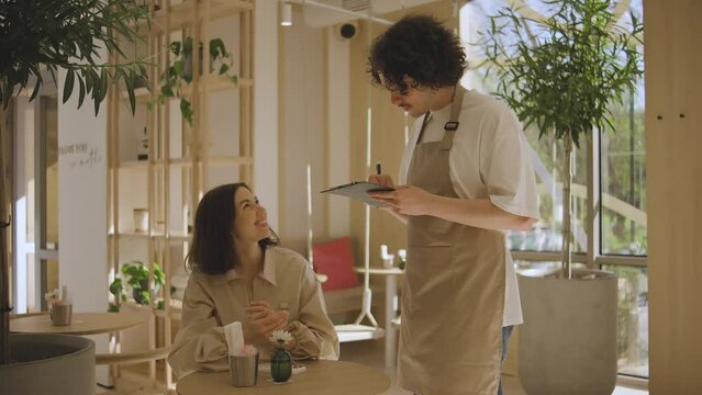 A waiter takes an order from a girl who is sitting at a table in a cafe. A beautiful girl in a good mood communicates with a waiter