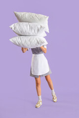 Female housekeeper with stack of white pillows on lilac background