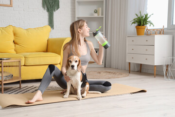 Sporty young woman with Beagle dog drinking water on yoga mat at home