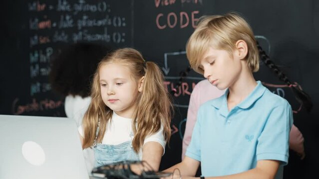 Smart girl using laptop programing engineering code with diverse friends ay table with microchip and main board scatter around in STEM classroom at blackboard written with coding prompt. Erudition.