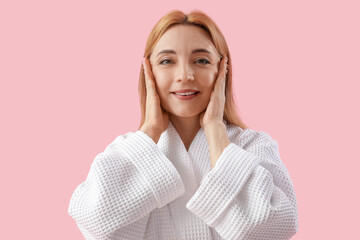 Beautiful happy mature woman in bathrobe doing face building exercise on pink background