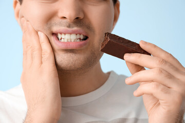 Young man with candy bar suffering from toothache on blue background, closeup