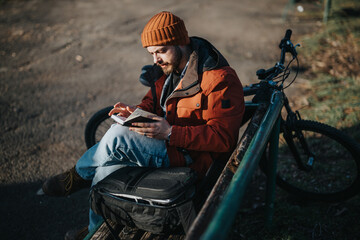 A relaxed young man with facial hair in casual attire spends leisure time reading outdoors, with...