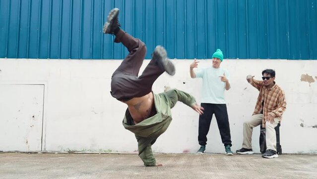 Skilled hipster doing cart wheel while multicultural friends encourage him. Hip-hop dancer moving foot step to music surrounded by group of hipster at wall near street. Outdoor sport 2024. hiphop.