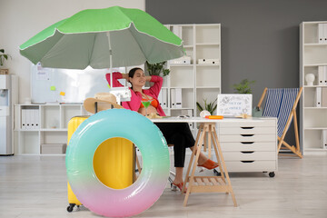 Happy female office worker sitting at table near suitcase and inflatable ring in office. Summer...