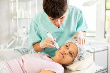 Young male cosmetologist performs hardware facial microcurrent procedure to young female patient
