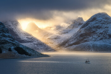 Snowy mountains in clouds, bright sunbeams, boat, sea at sunset