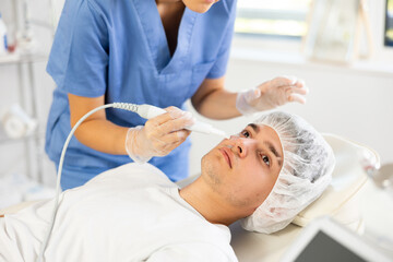 Young female cosmetologist performs hardware facial exfoliation procedure to young male patient