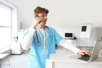 Male medical intern with laptop talking by mobile phone at reception in clinic