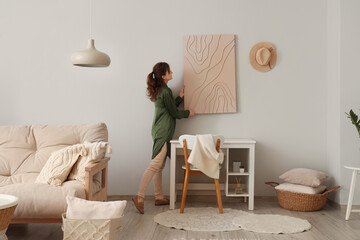 Young woman hanging picture in stylish living room - obrazy, fototapety, plakaty