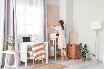 Young pretty woman hanging picture on wall near workplace in room