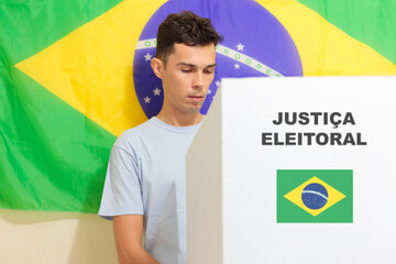 Jovem homem brasileiro escolhendo o seu candidato na urna eletrônica. Na parede, a bandeira do Brasil decora o local de votação. A imagem retrata as eleições brasileiras.