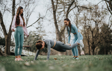 Two athletic women engage in a fitness routine in a serene park setting, demonstrating strength and flexibility.
