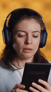 Vertical video Woman wearing headphones, practicing new language using internet app on digital tablet, isolated over studio background. Person repeating foreign words, reading them on device