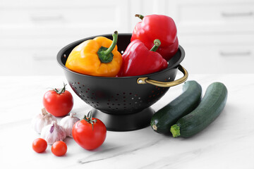 Black colander and different vegetables on white marble table, closeup