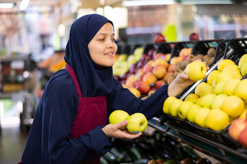 Young woman seller in hijb and apron sells apples in grocery store