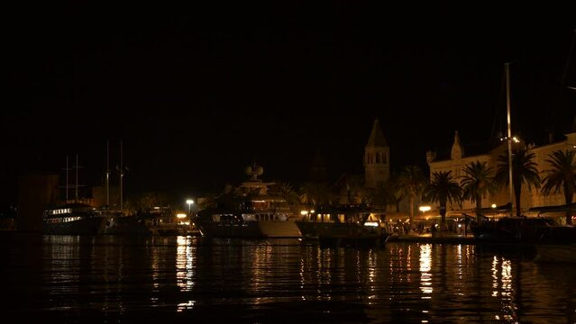 Lovely seaside town in Croatia with street lights reflecting in calm harbour sea and smaller boats sail passing large vessels moored along the promenade. Quiet evening in old city harbour of Trogir.